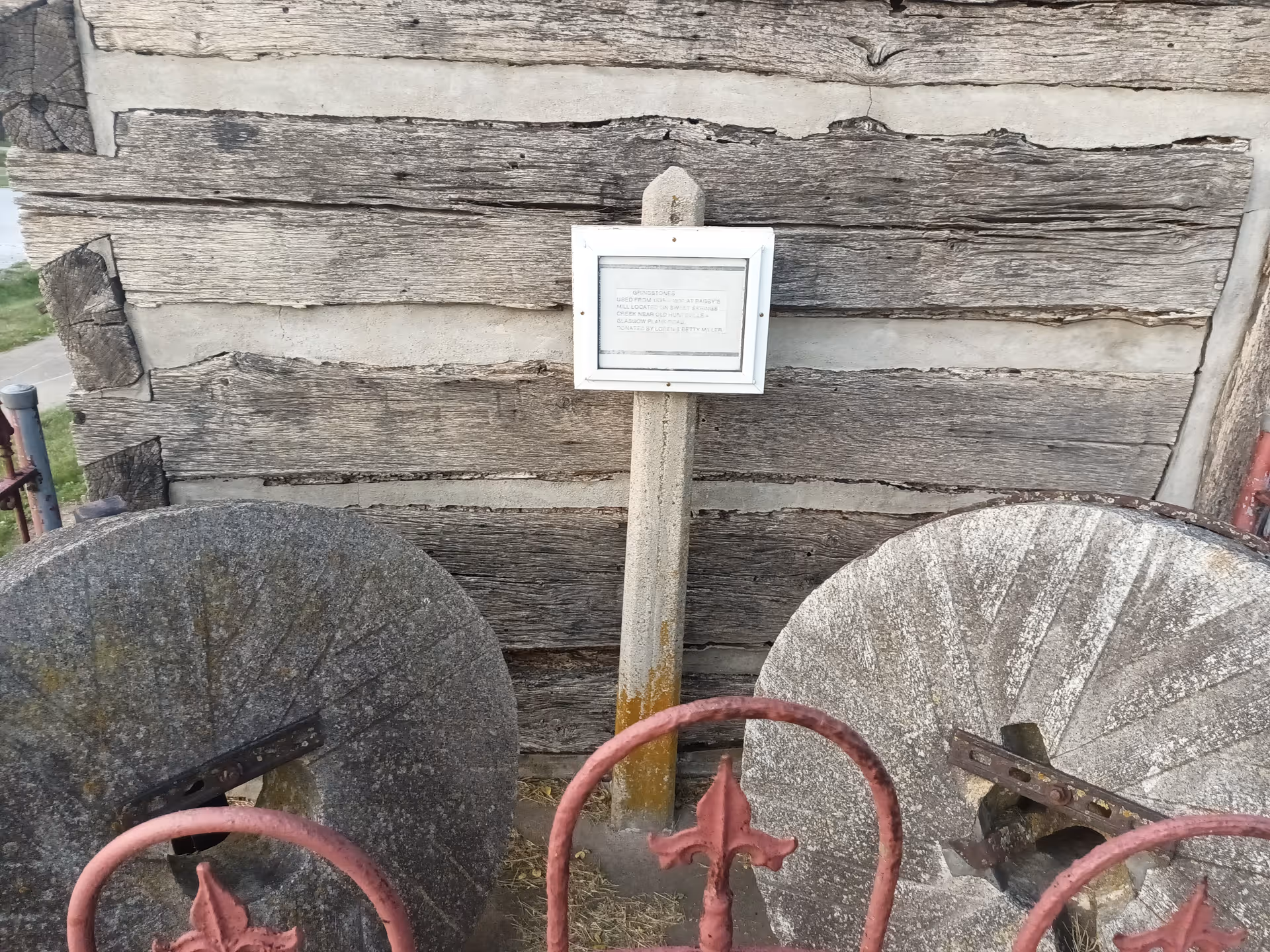 Weathered wooden building exterior with a sign, two stone mill wheels, and red metal rails.