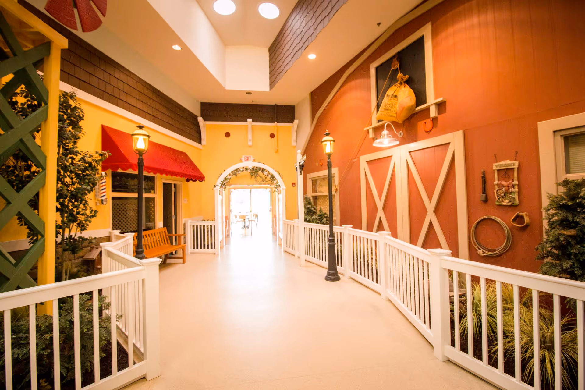 Indoor hallway designed to resemble an outdoor street scene with white railings, lamp posts, a wooden bench, and walls painted to look like building facades including a red barn door and a storefront with a red awning.
