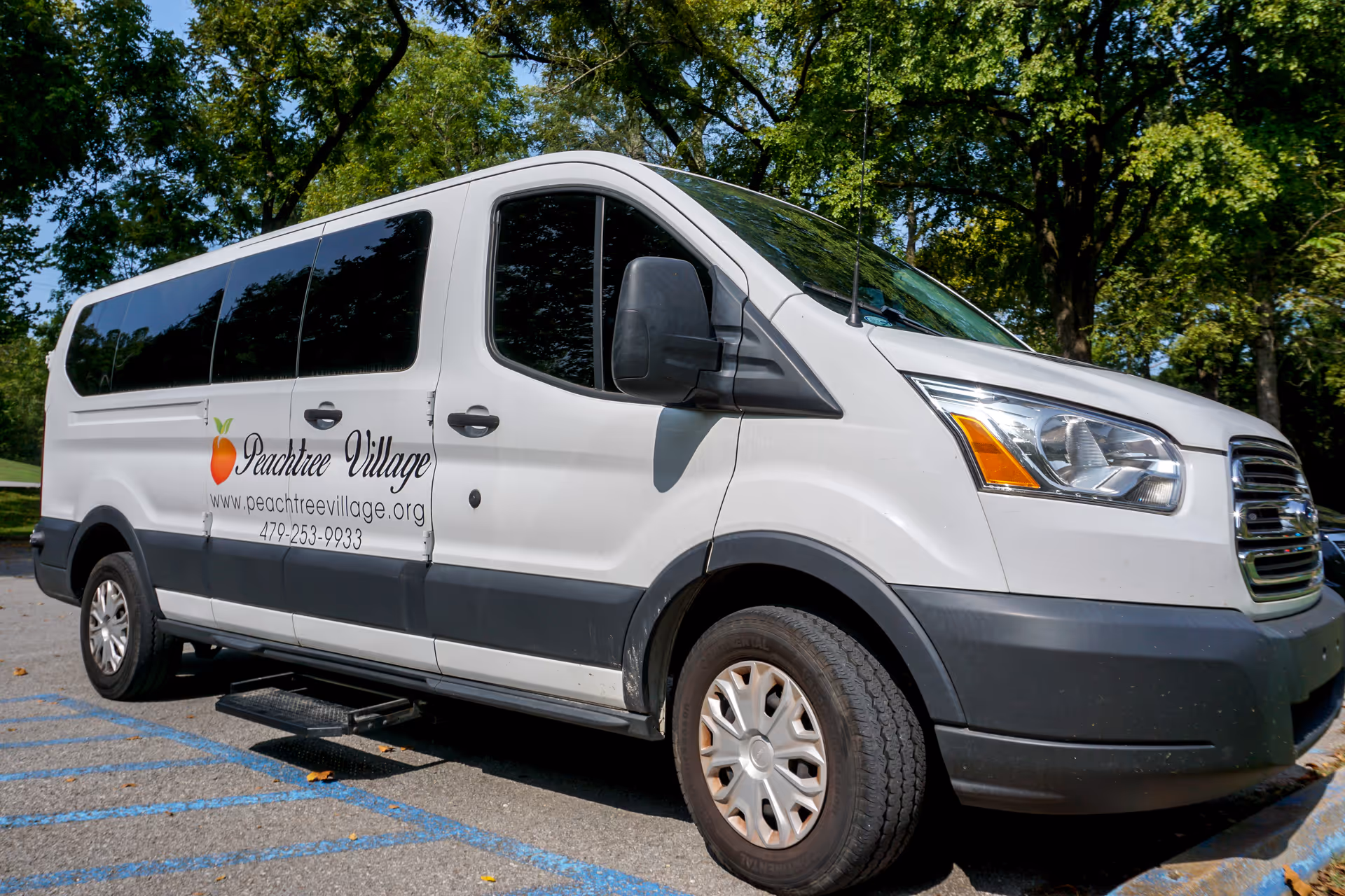 A white Peachtree Village shuttle van parked under trees with the facility logo and contact information on its side.