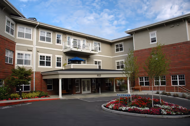 Multi-story senior living facility with a covered entrance, balcony, and landscaped flower bed.