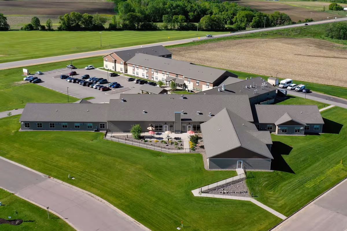 Aerial view of Dove Healthcare – Osseo Assisted Living facility showing a large building complex with multiple wings surrounded by well-maintained green lawns, parking lots with several cars, and adjacent farmland.