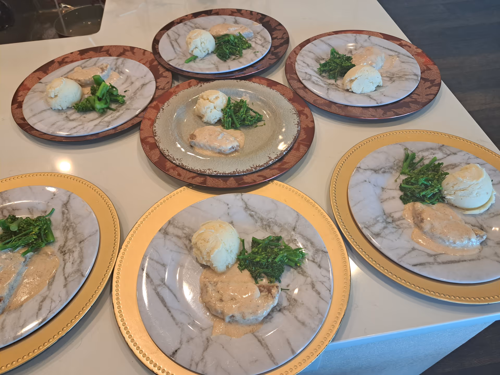 Multiple marble plates with plated meals, greens, and sauce on a white table.