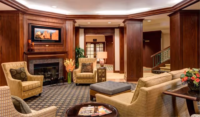 A cozy living room area with wood-paneled walls and columns, featuring a fireplace with a framed picture above it. The room has comfortable beige armchairs and sofas with patterned cushions, a gray ottoman, a round wooden coffee table with magazines, and a side table with a lamp and a vase of flowers. There is a staircase visible in the background and warm lighting throughout the space.