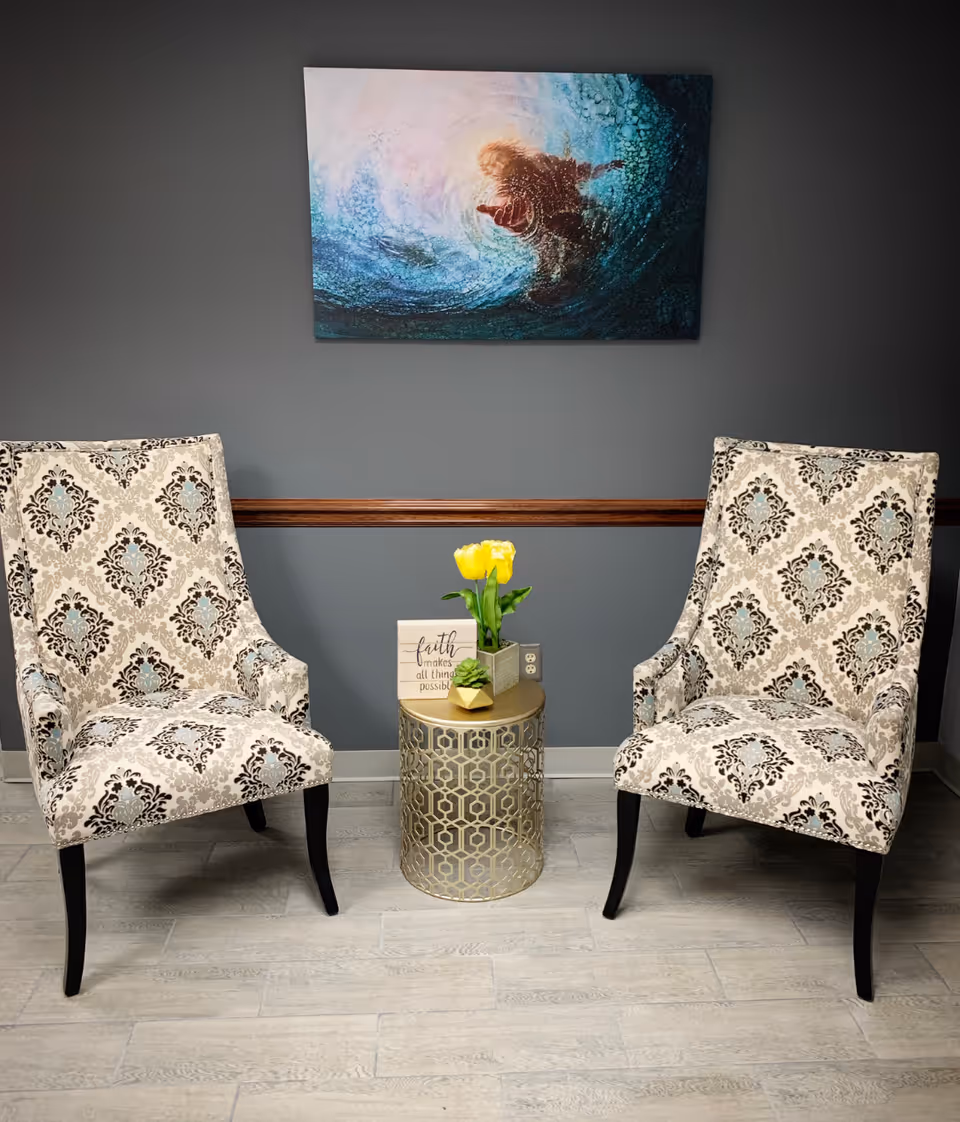Two patterned chairs flank a gold lattice table with yellow tulips against a gray wall.