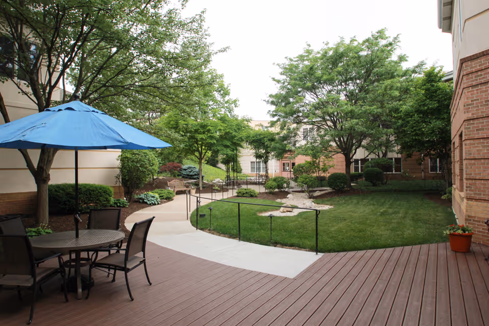 Courtyard patio with a table and blue umbrella overlooking a landscaped lawn, trees, and surrounding building.