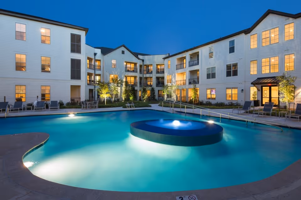 Evening view of a well-lit outdoor swimming pool surrounded by a three-story white residential building with balconies and large windows. The pool area has lounge chairs and tables, with some greenery and trees around the building.