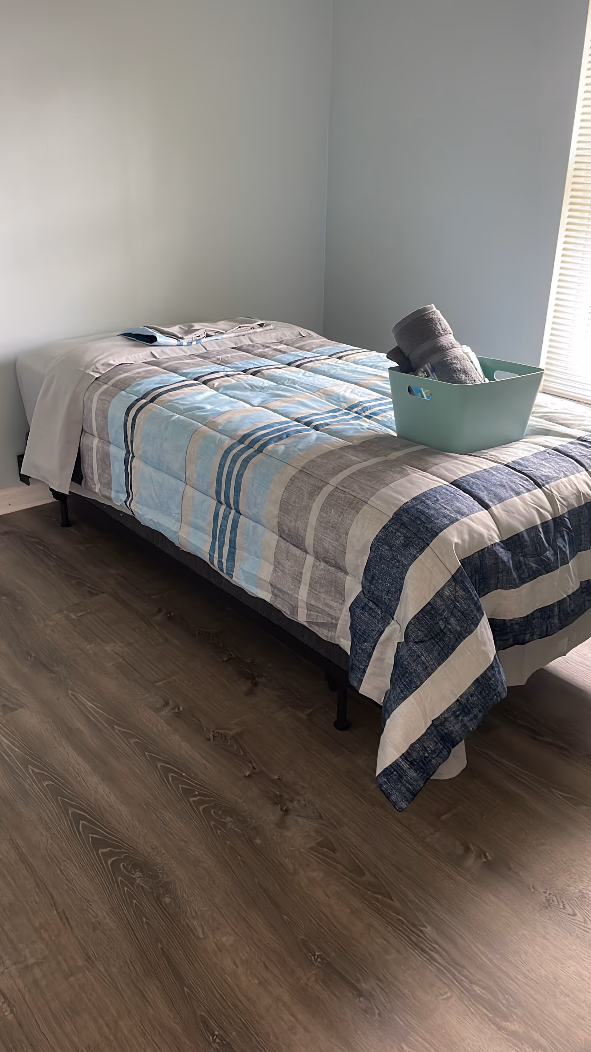 A bedroom with a striped quilt, light walls, and a plastic basket on the bed.