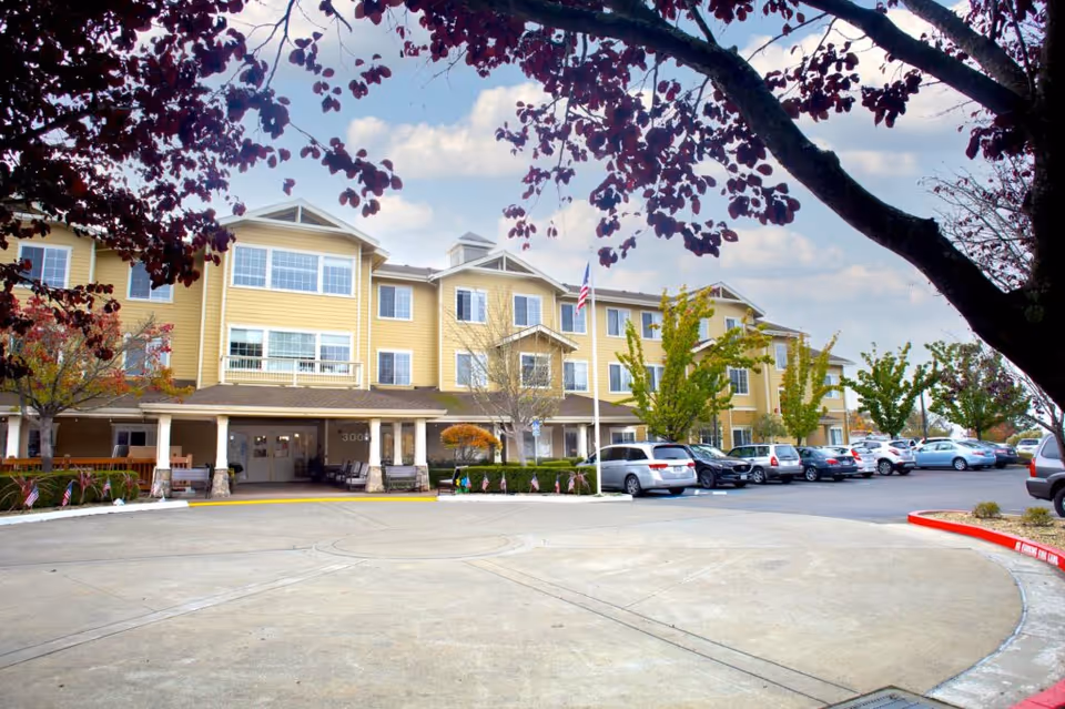 Front exterior view of a three-story yellow senior living facility building with multiple windows, a covered entrance, several parked cars in the parking lot, and trees with autumn foliage framing the scene.