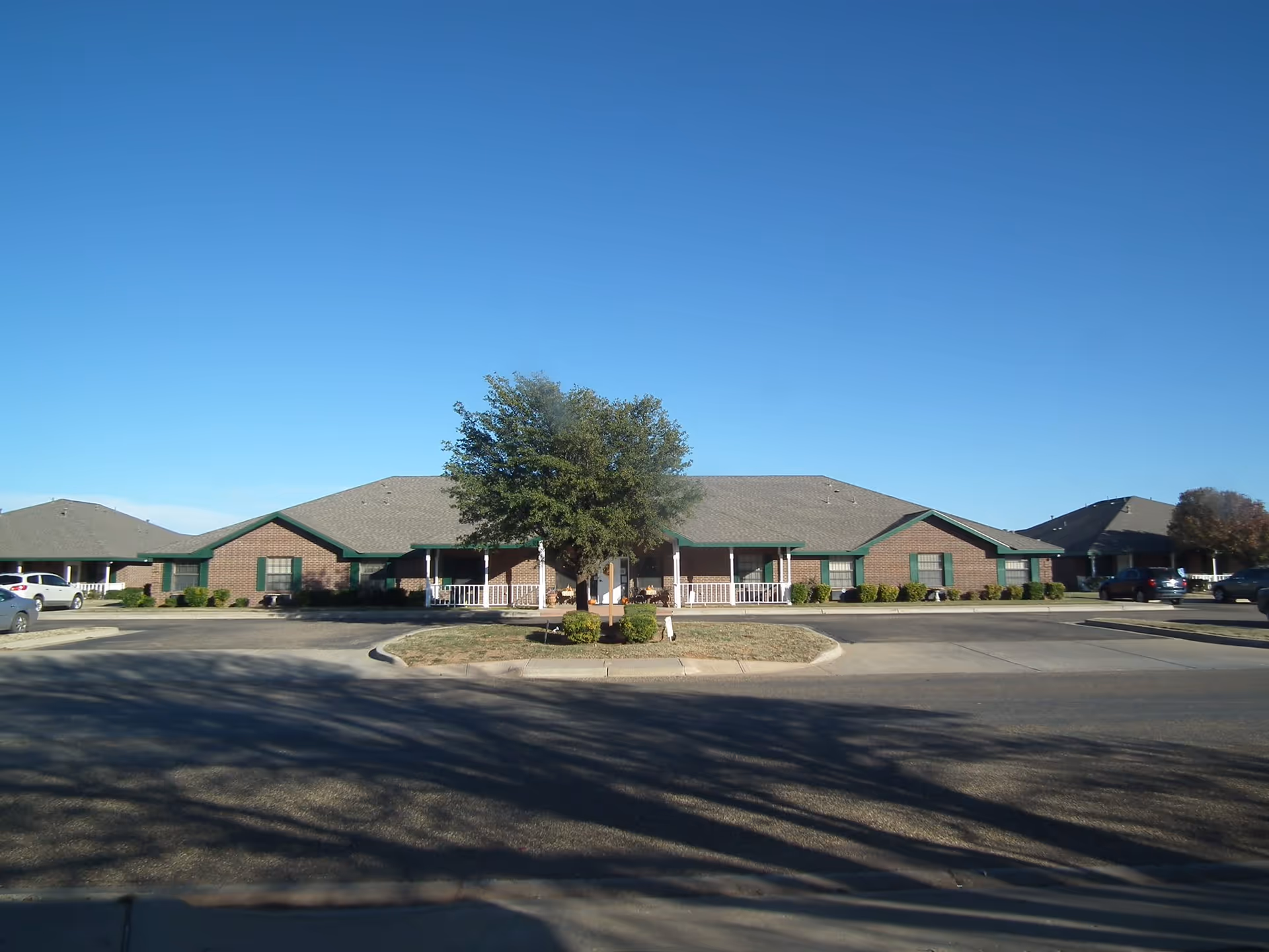 One-story brick senior living building with a central porch and tree in a circular driveway under a clear blue sky.