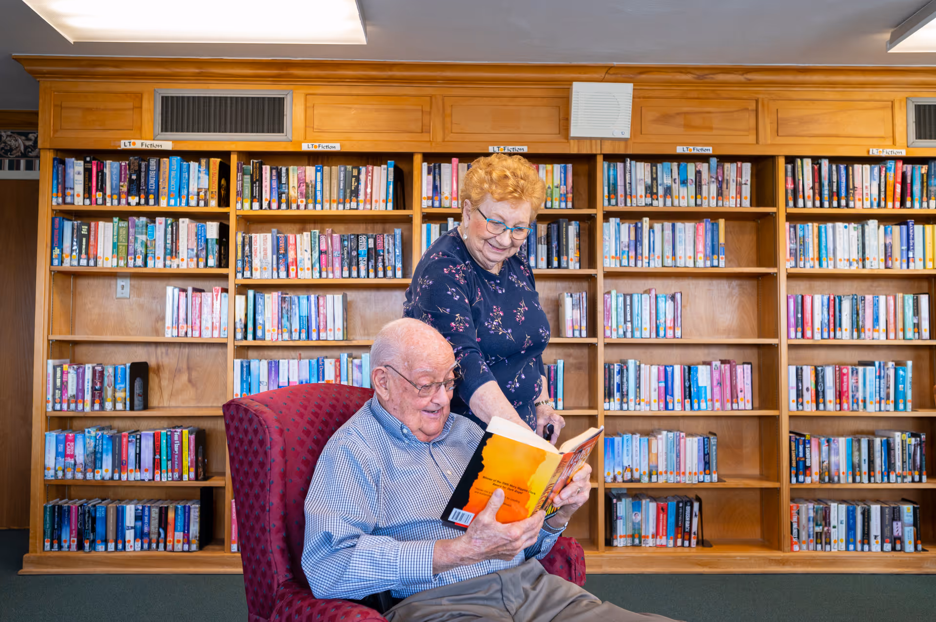An elderly man sitting in a red armchair reading a book while an elderly woman stands beside him, smiling and looking at the book. They are in a library with wooden bookshelves filled with books in the background.