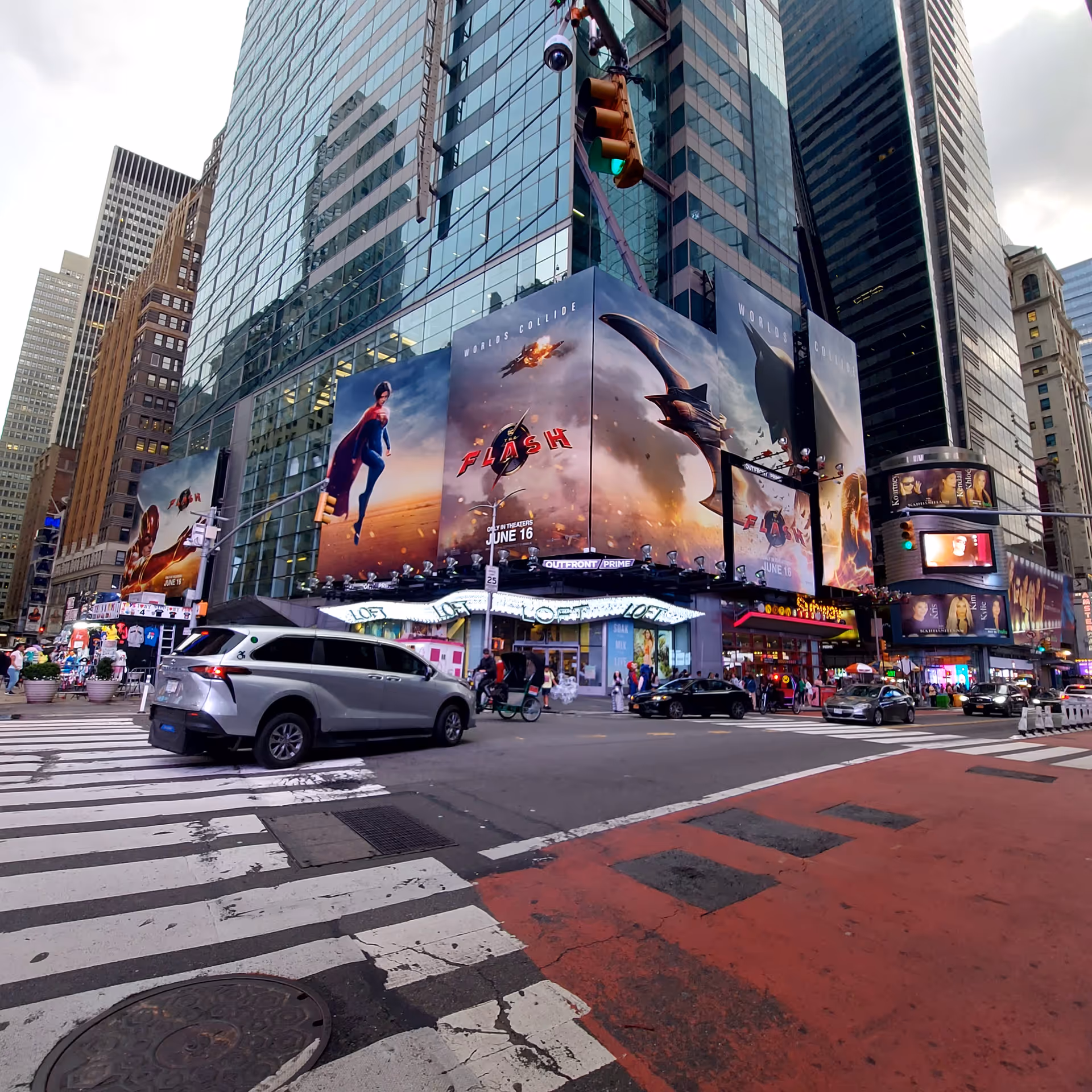 Busy city intersection with towering glass buildings and large movie billboards, traffic and pedestrians.