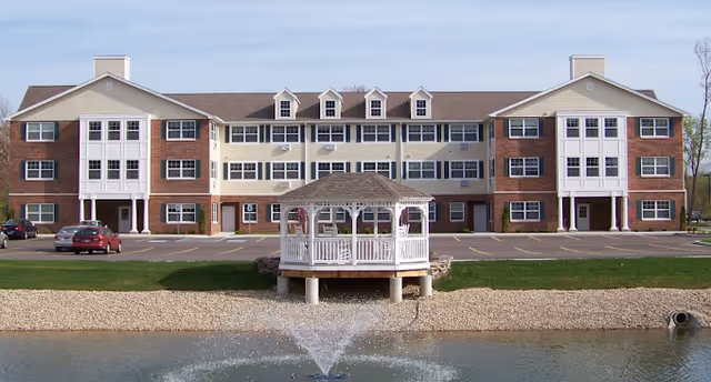 Front view of a three-story brick-and-siding senior living building with a white gazebo and a fountain in a pond in the foreground.