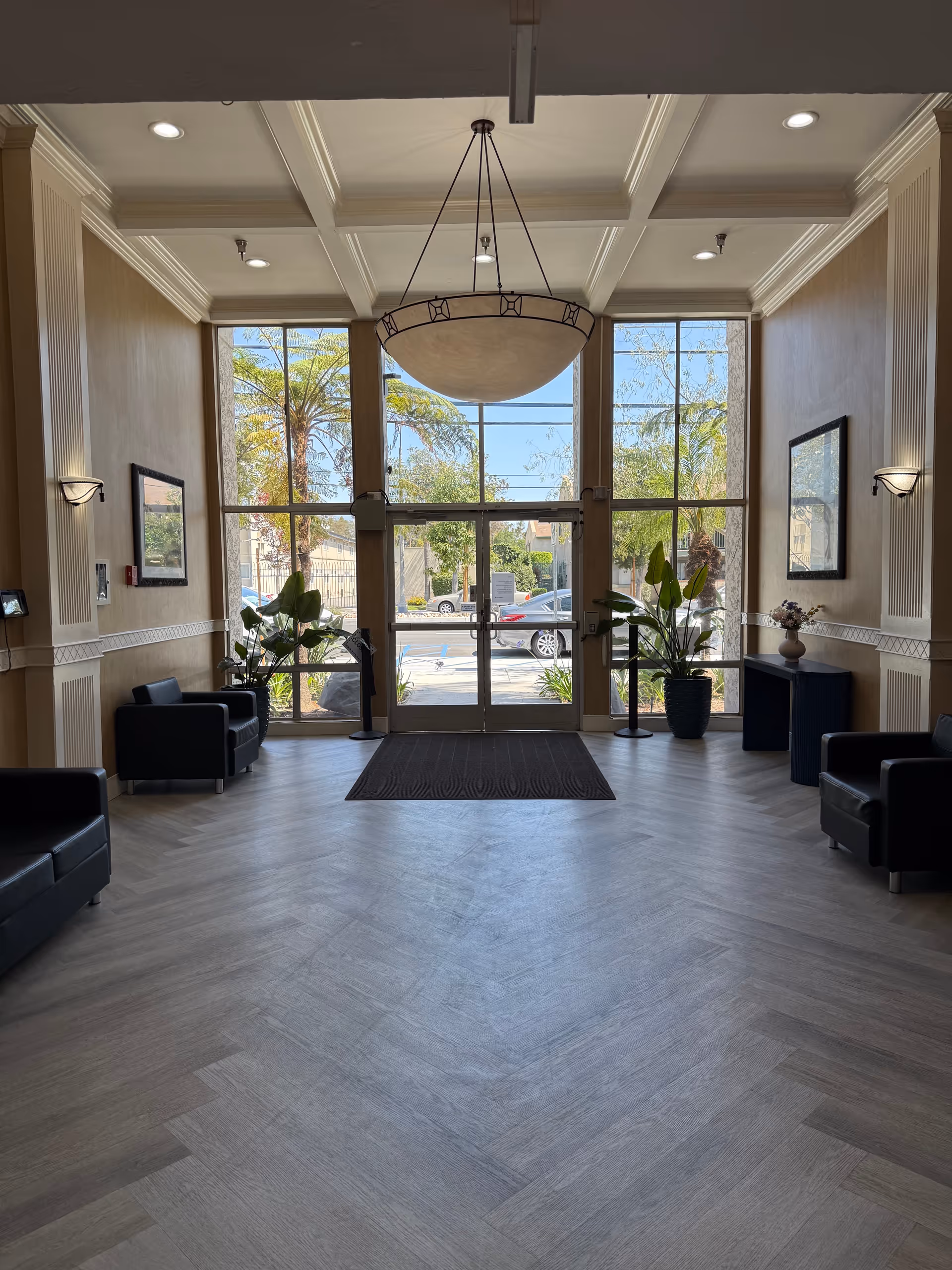 Bright lobby with glass double doors, large circular chandelier, plants, and black leather seating.