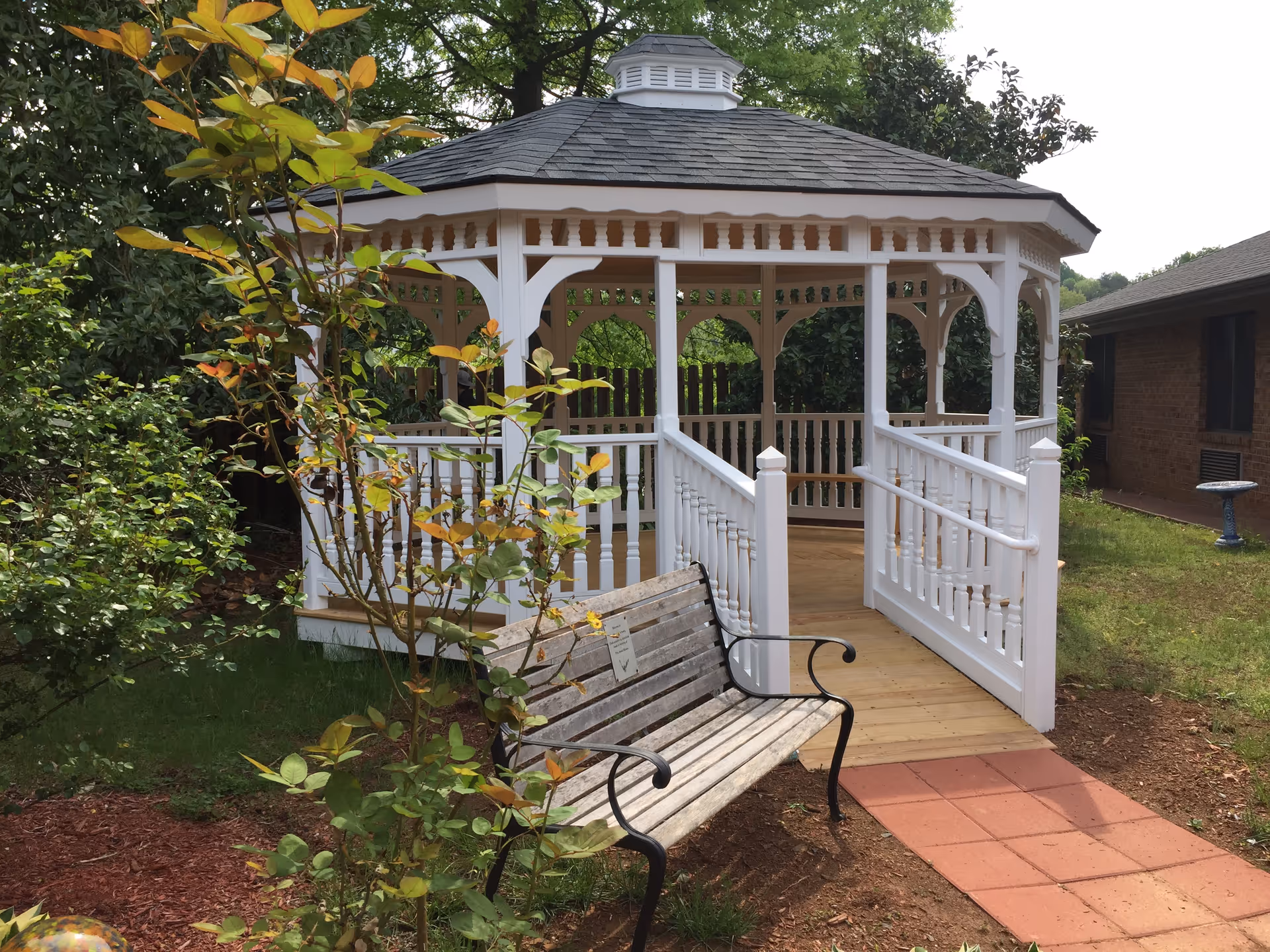 A white wooden gazebo with a shingled roof and decorative railings in a garden area. There is a wooden bench with black metal armrests in front of the gazebo, surrounded by green bushes and trees. A brick building and a birdbath are visible in the background.