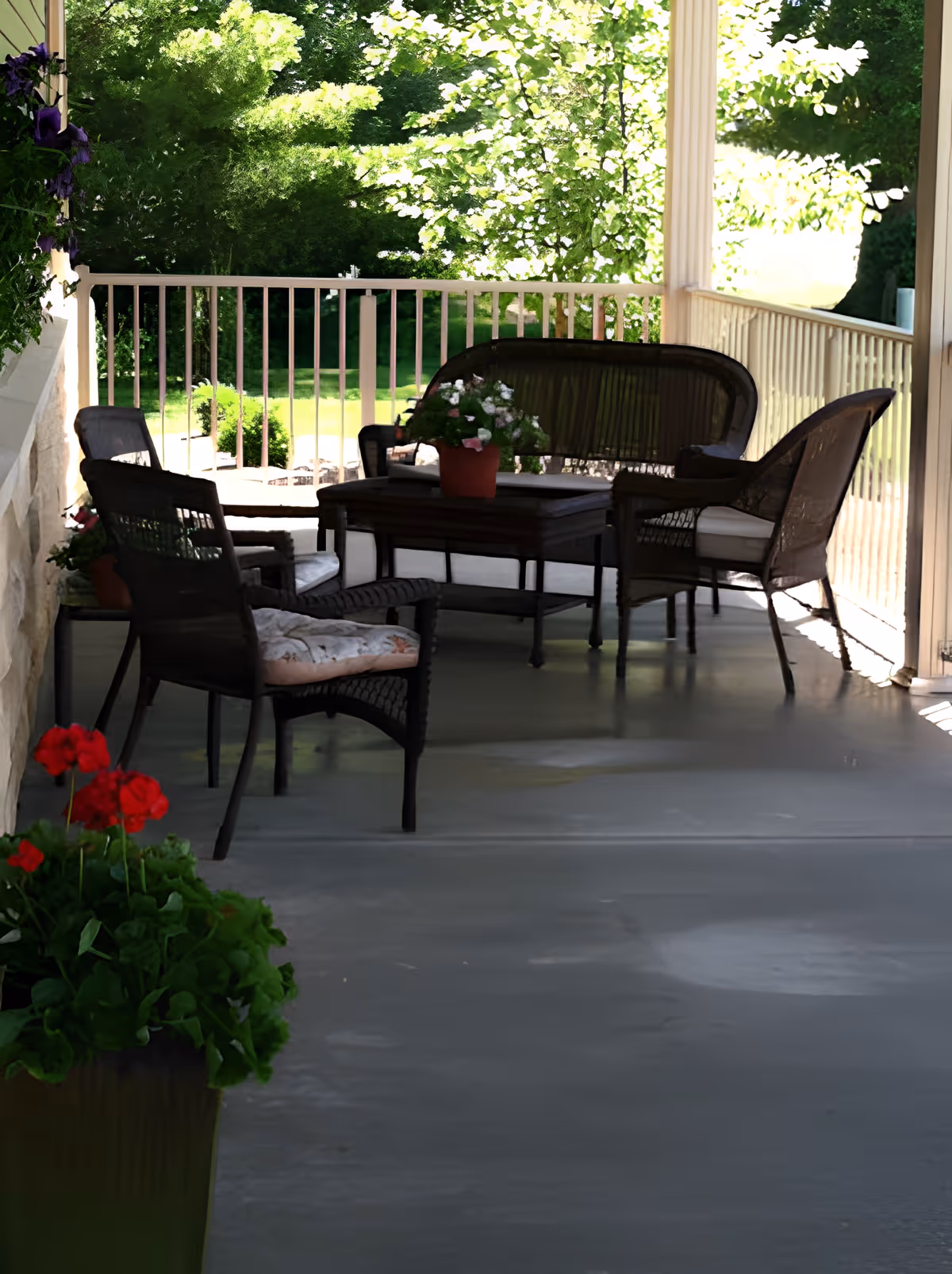 A covered outdoor porch area with wicker chairs and a loveseat arranged around a wooden coffee table with a potted plant on top. The porch has a railing and overlooks a green garden with trees and sunlight filtering through the leaves. There are also potted flowers placed on the porch floor and hanging on the wall.