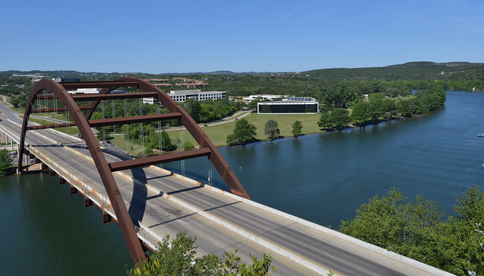 Aerial view of a curved brown arch bridge over a river with trees and campus buildings.