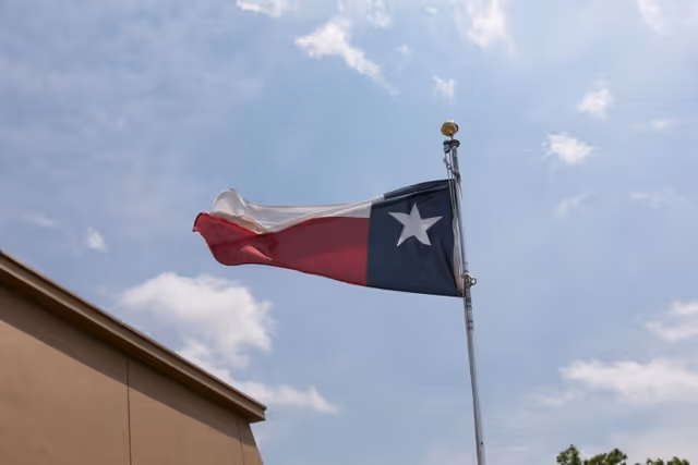A Texas state flag waving on a flagpole against a partly cloudy sky, with the corner of a building visible at the bottom left.