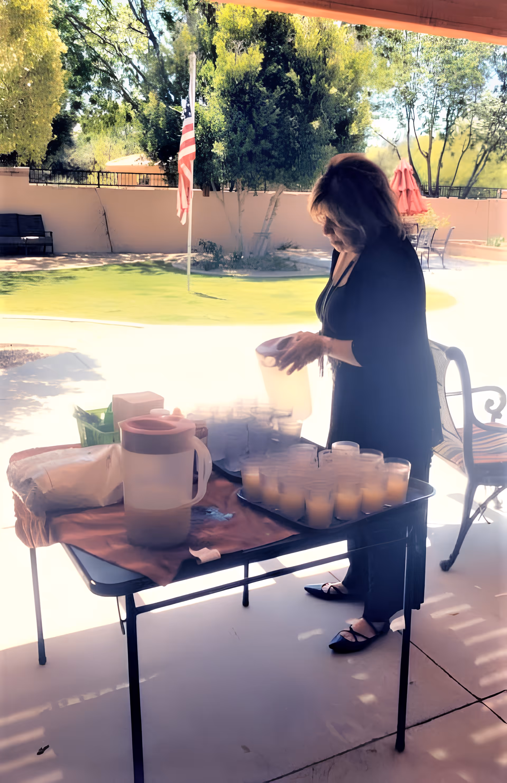 A woman standing outdoors under a shaded patio area, pouring drinks into plastic cups arranged on a table. The background shows a green lawn, trees, an American flag on a flagpole, and outdoor seating with umbrellas.