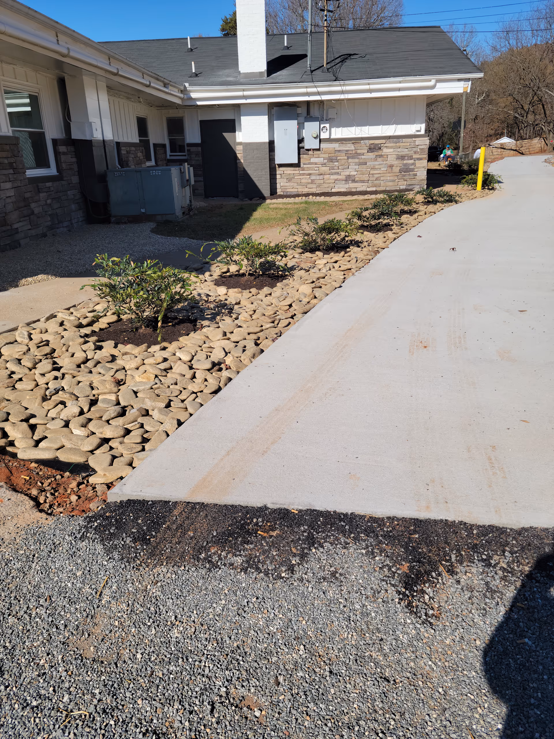 Concrete driveway and landscaped rockbed with small shrubs alongside the exterior wall of a single-story building.