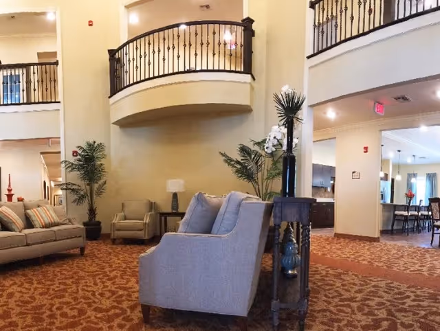 A spacious living room area in a senior living facility with beige walls and patterned carpet. The room features several upholstered chairs and sofas arranged for conversation, potted plants, a small side table with a lamp, and a decorative railing on the upper level overlooking the space. In the background, there is a dining area with tables and chairs visible through an open doorway.