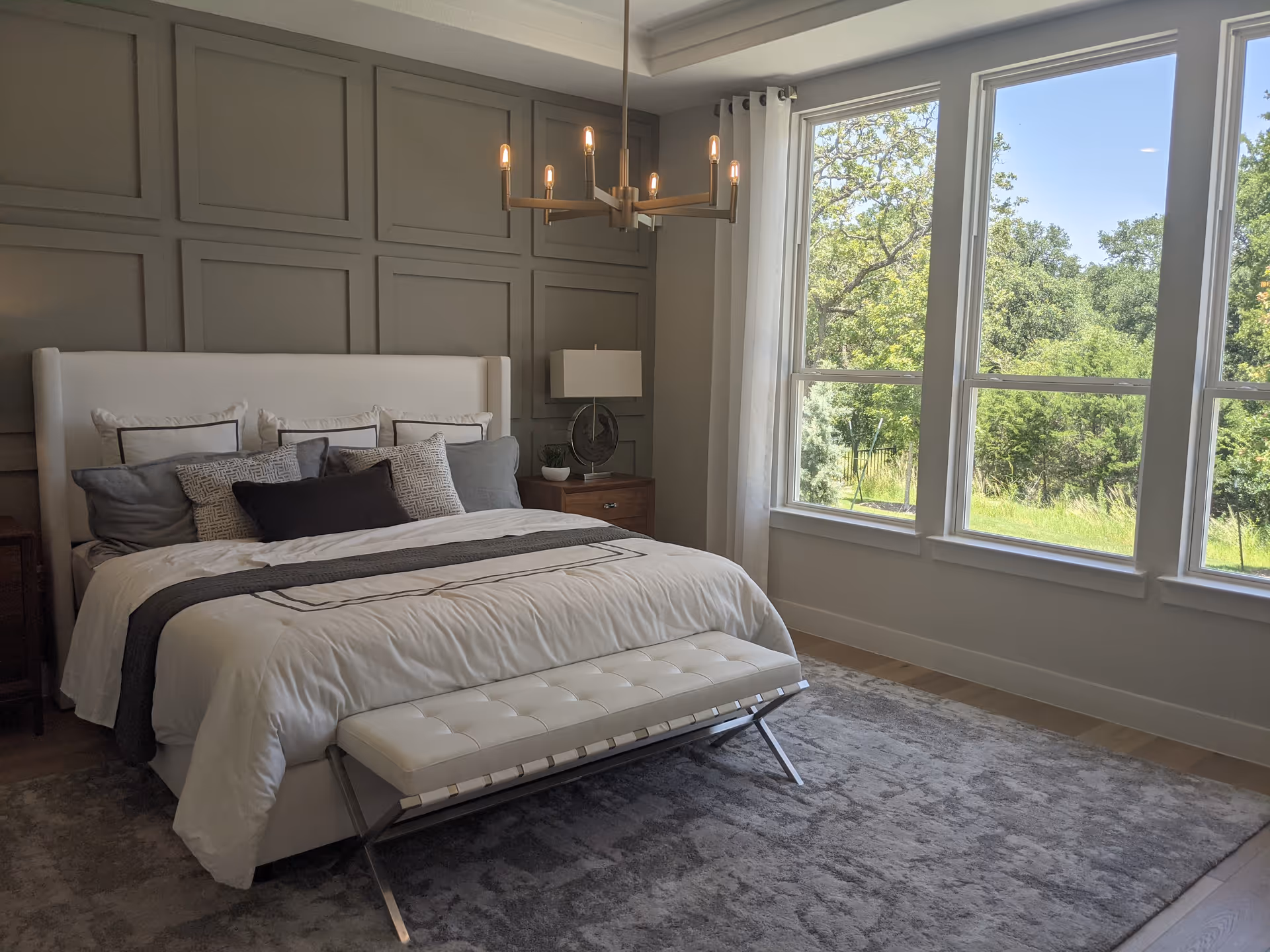 Sunlit bedroom with gray paneled wall, king-size bed, bench at foot, and large windows.