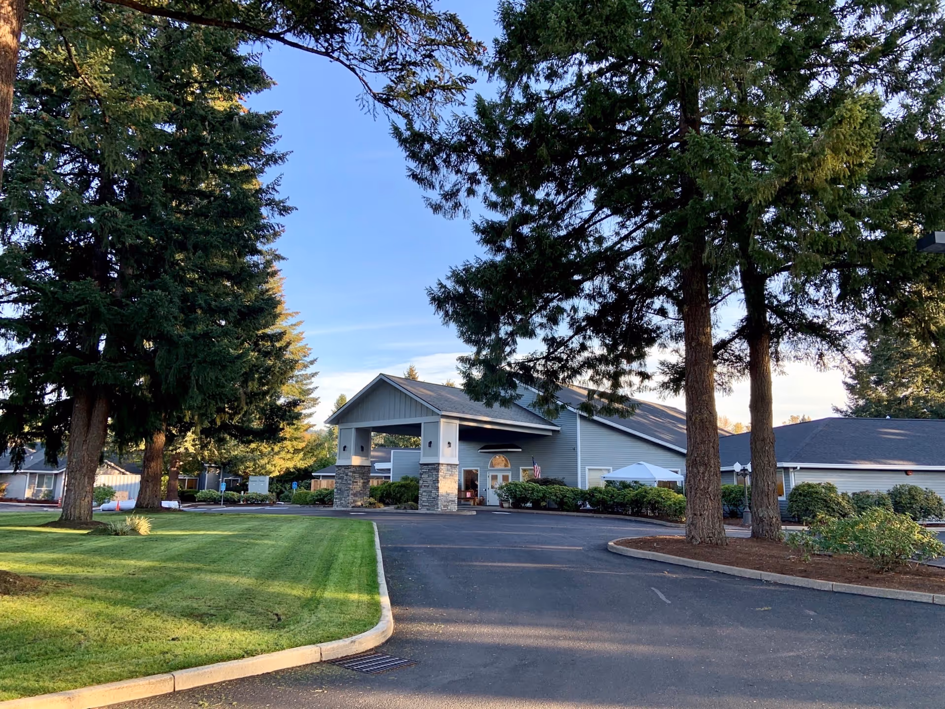Front exterior of a single-story senior living building with a covered entrance, driveway, and tall trees on a manicured lawn.