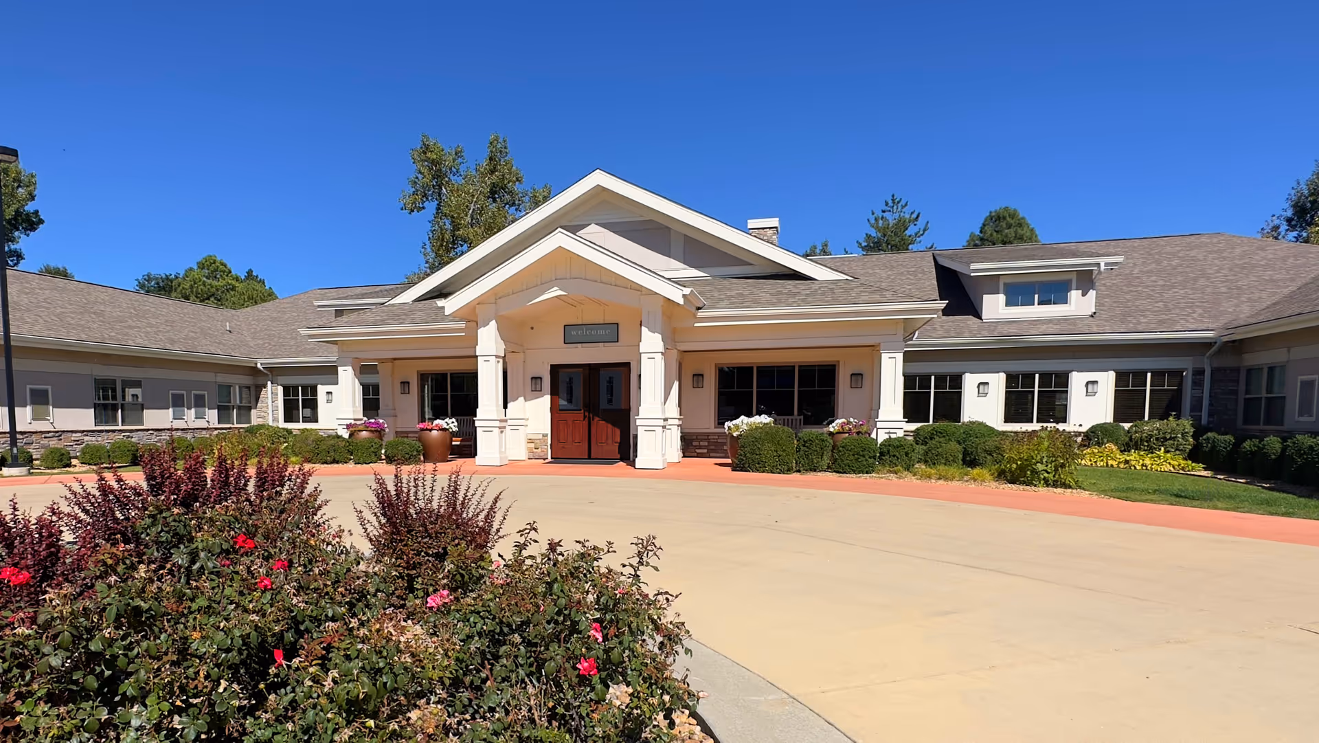 Front entrance of a single-story memory care building with a covered portico, red double doors, landscaped flower beds, and clear blue sky.