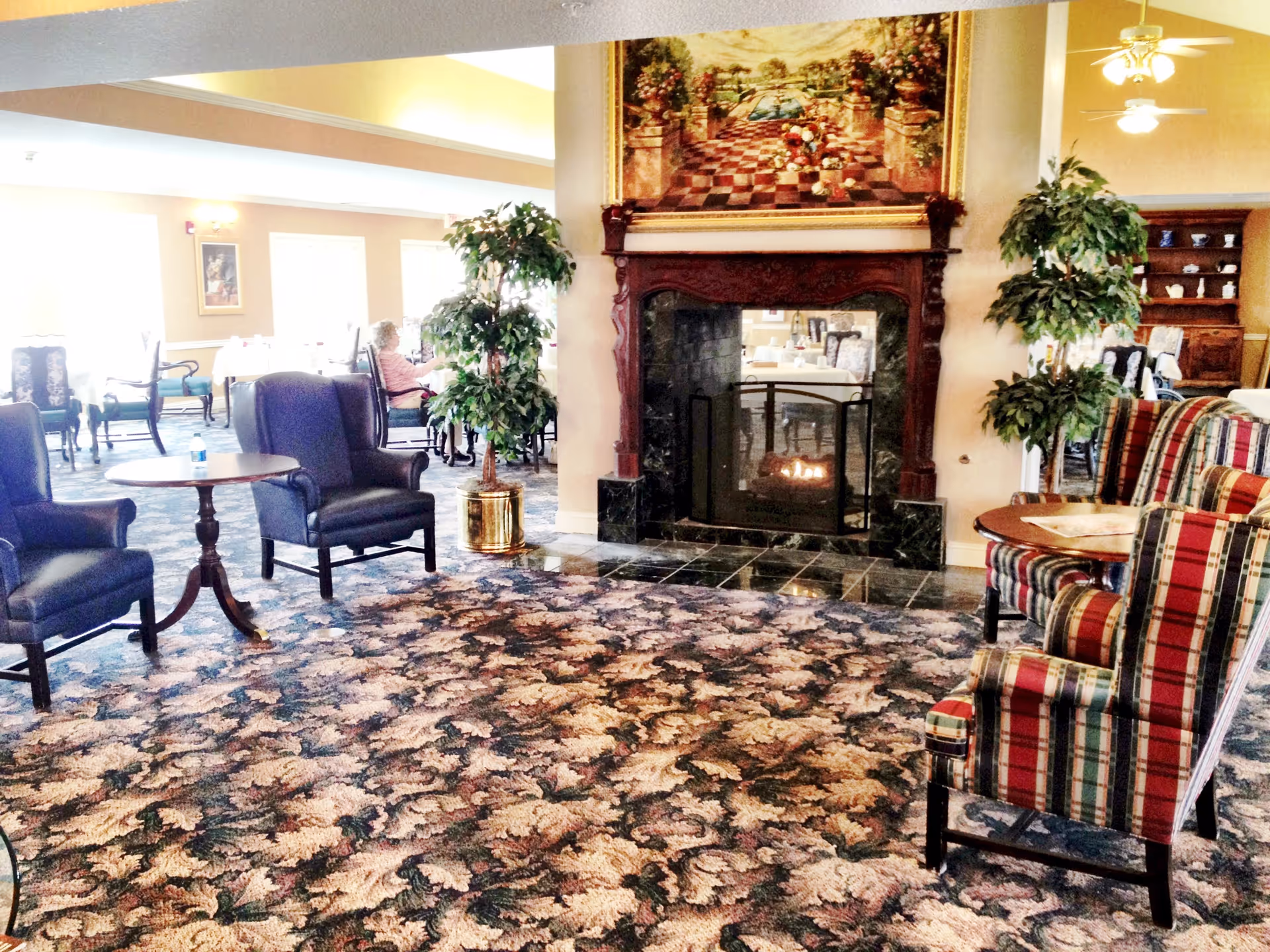 A bright senior living room with upholstered chairs and small tables arranged around a central fireplace and patterned carpet.