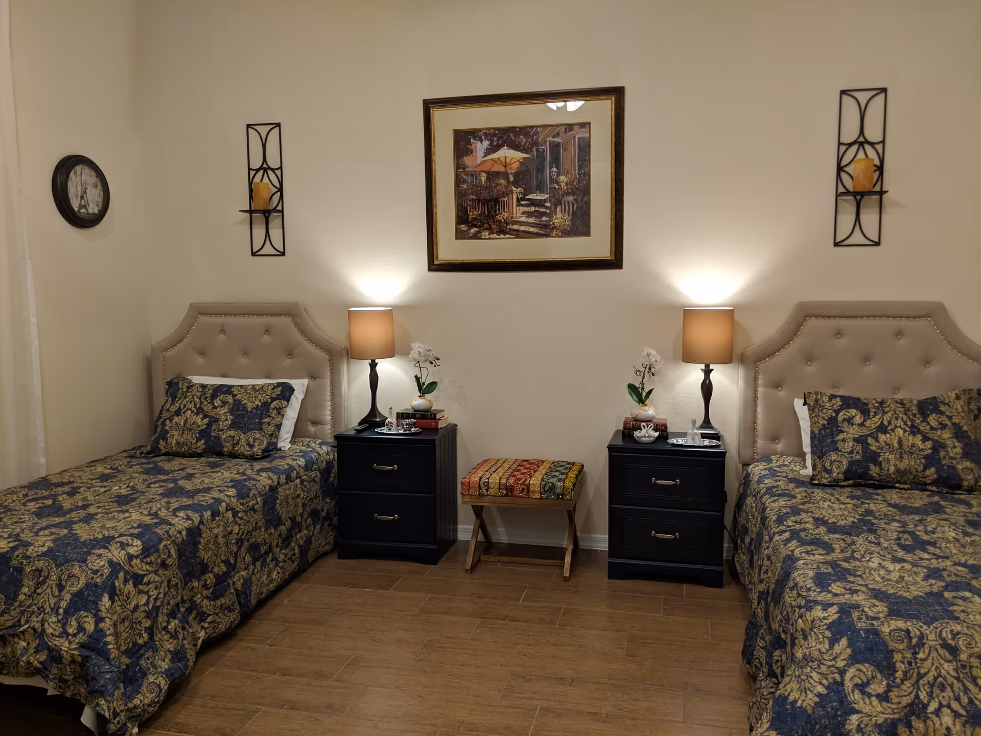 Symmetrical bedroom with two beige upholstered twin beds, nightstands, lamps, and a central bench.