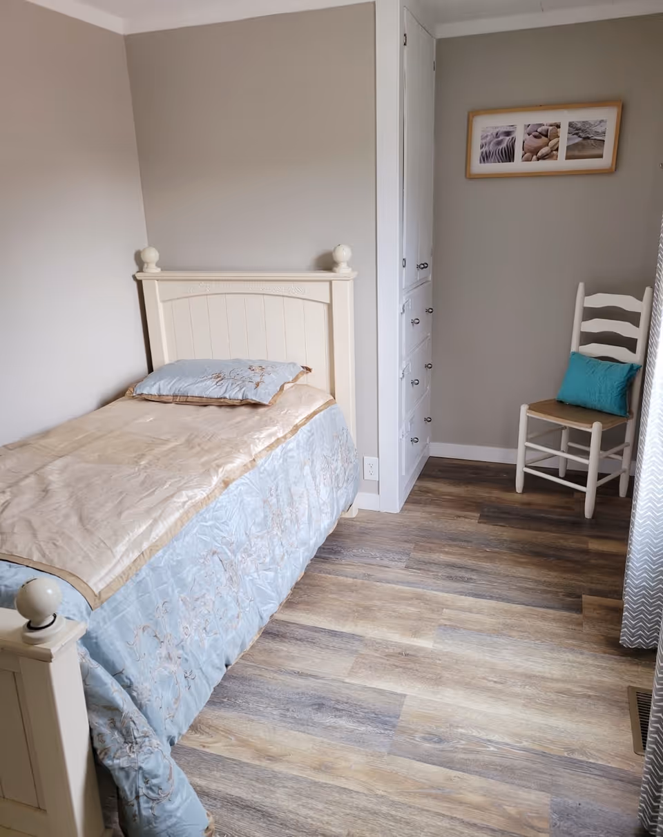 A small cream-colored bedroom with a single bed, dresser, chair, and framed art.
