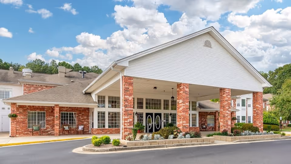 Exterior view of a senior living facility building with a large covered entrance supported by brick columns, surrounded by landscaped greenery and a clear blue sky with scattered clouds.