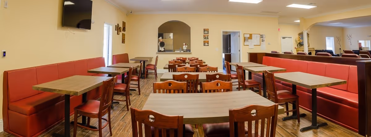 Interior view of a dining room in a senior living facility with multiple wooden tables and chairs arranged neatly. Red cushioned bench seating lines the walls, and there is a wall-mounted TV on the left side. The room has beige walls, wooden flooring, and a serving window to the kitchen area in the back.