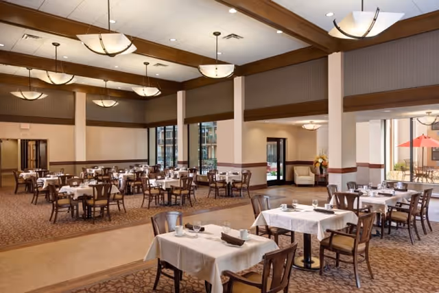 Large, well-lit dining room with tables covered in white linens, wooden chairs, and chandeliers.