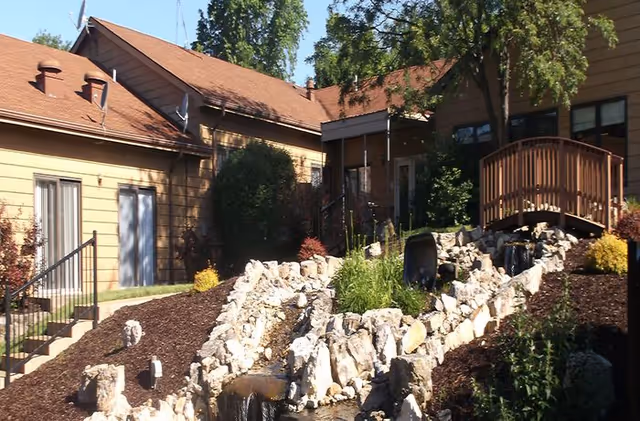Outdoor garden area at StoneBridge Senior Living - De Soto featuring a small wooden bridge over a rocky water feature with flowing water, surrounded by plants and shrubs, with the facility buildings in the background.