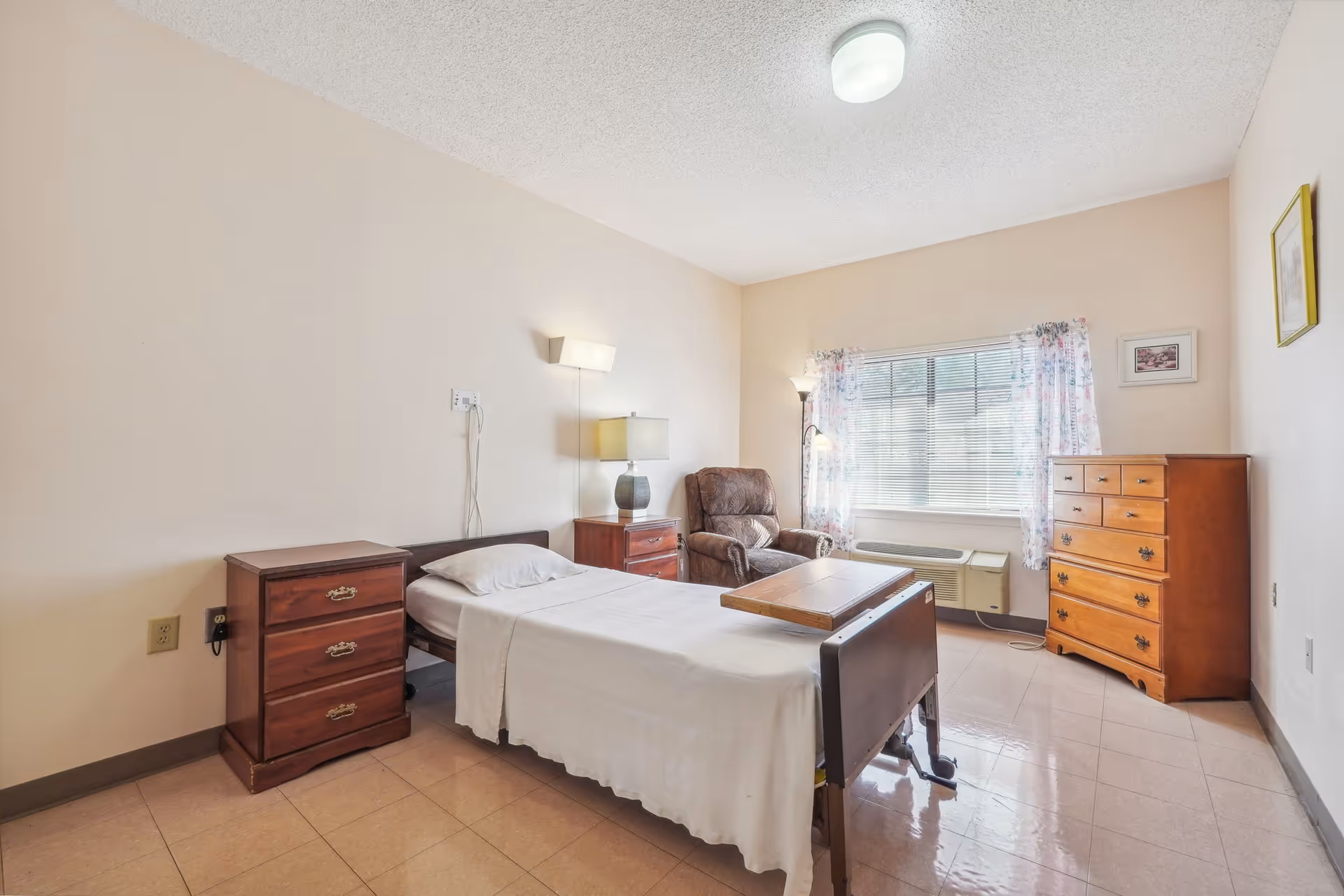 Bedroom in a senior living facility with a bed, dressers, chair, and window with curtains.