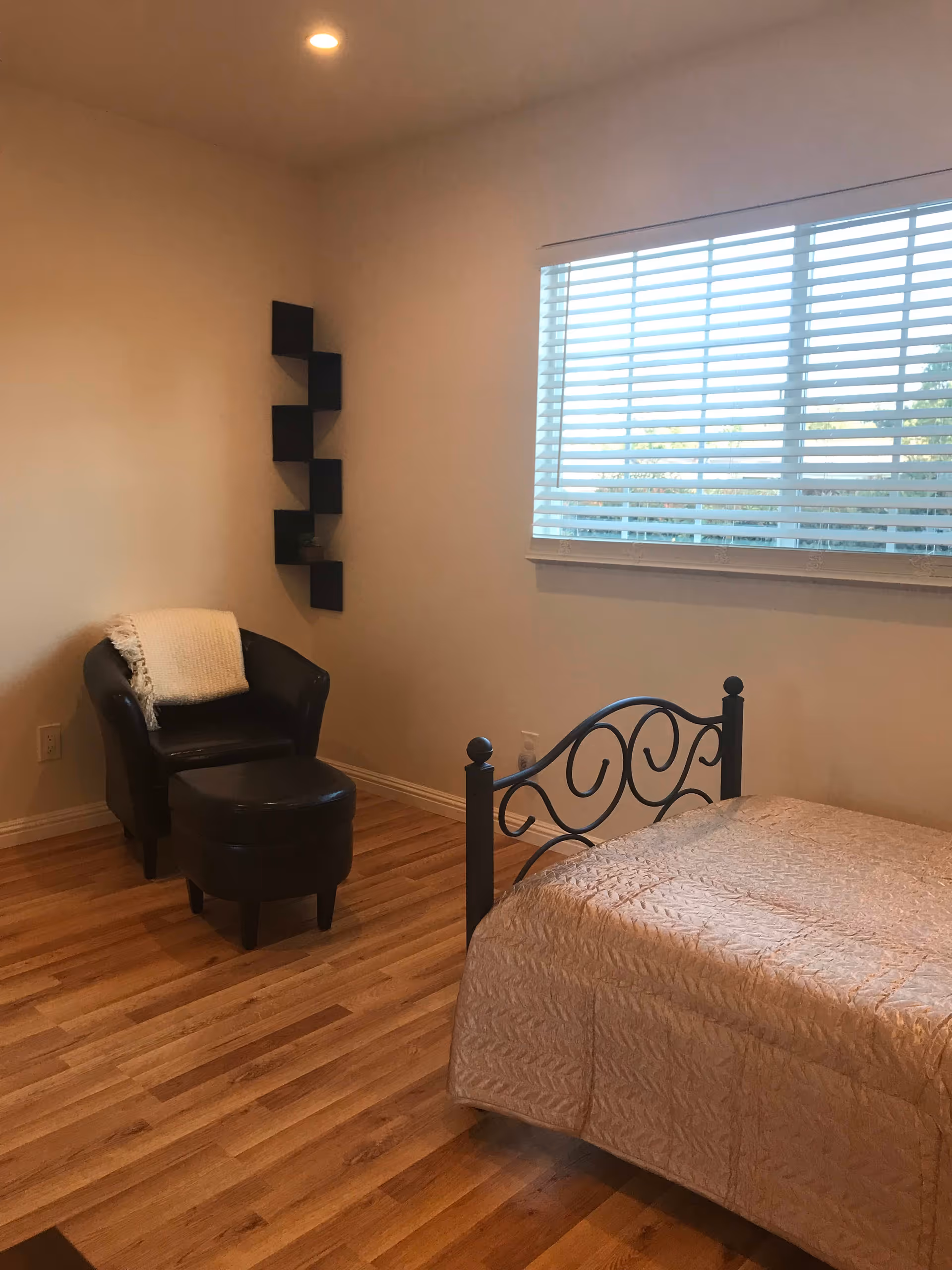 Simple bedroom with a metal-framed bed and quilted bedspread, a leather armchair with ottoman, corner wall shelves, and a window with blinds over wood floors.