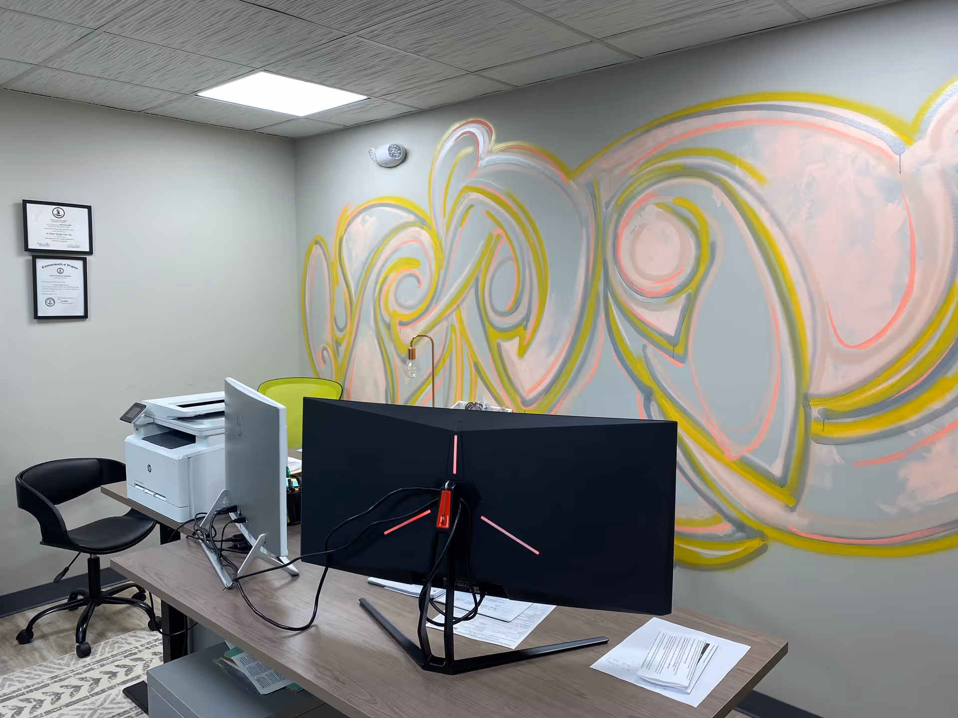 Office desk with computer monitor, printer, and colorful mural on the wall.