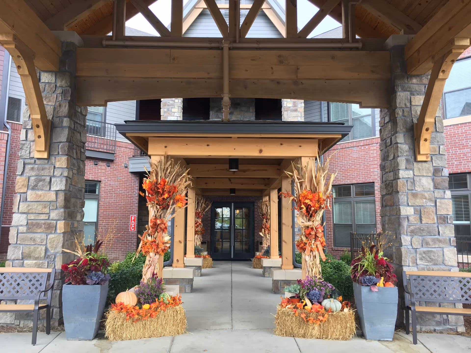 Covered entrance to a building with stone pillars and wooden beams, decorated with autumn-themed arrangements including hay bales, pumpkins, orange leaves, and dried corn stalks. Two benches and large planters with plants flank the walkway leading to glass doors.