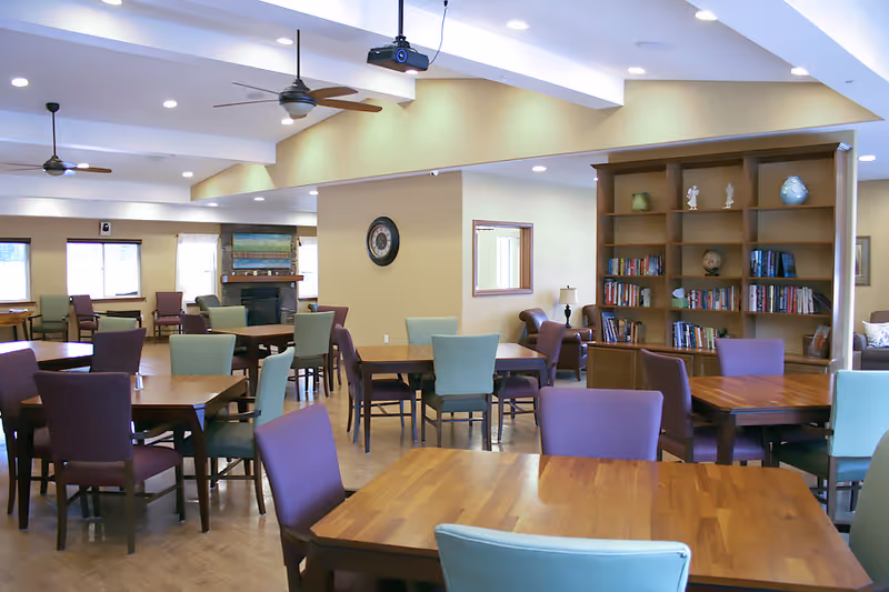 A bright communal dining area with wooden tables, colorful chairs, and a tall bookshelf.