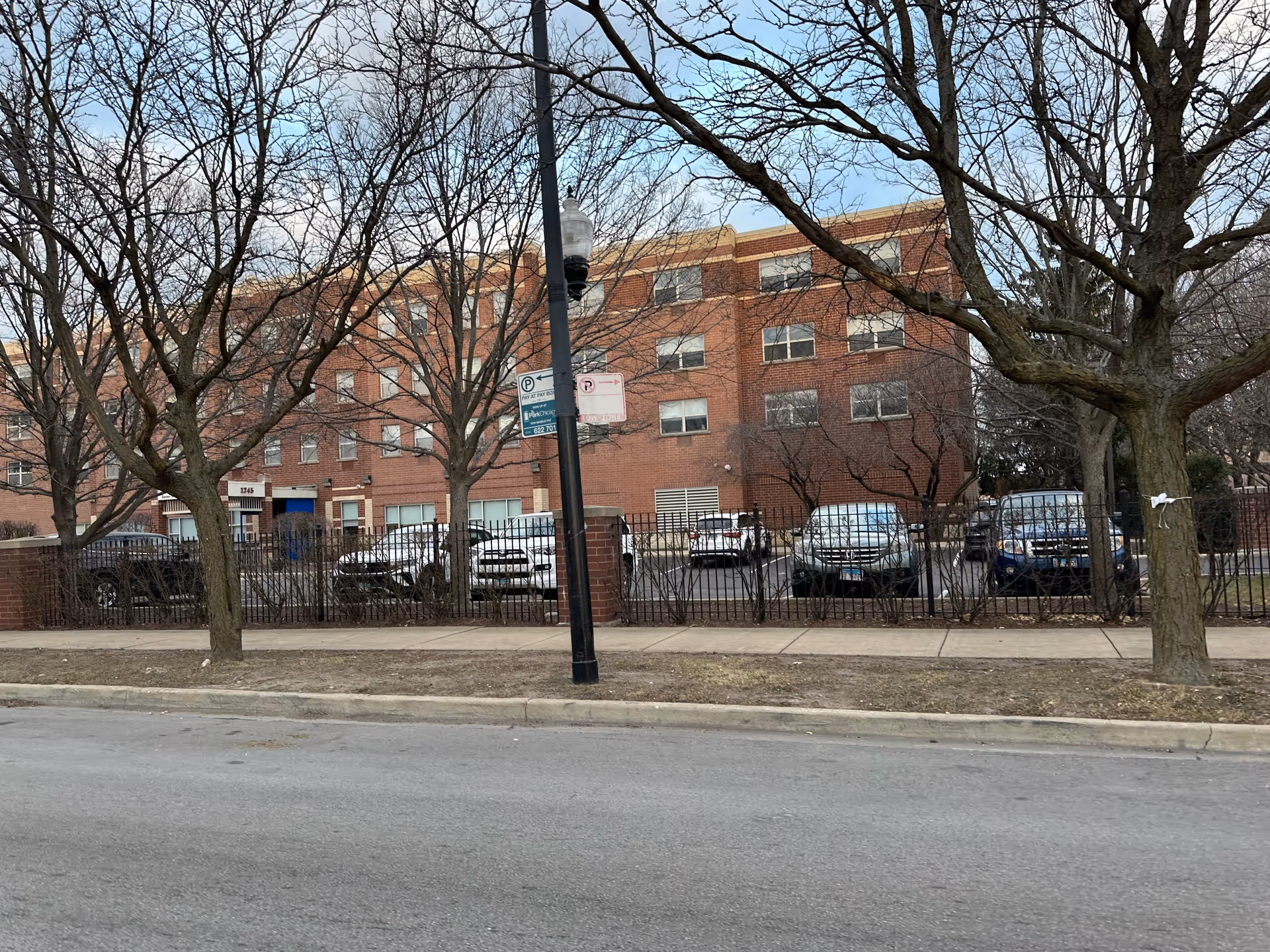 Exterior view of a multi-story brick building behind a black metal fence with several parked cars and leafless trees in front. A sidewalk and street are visible in the foreground under a partly cloudy sky.