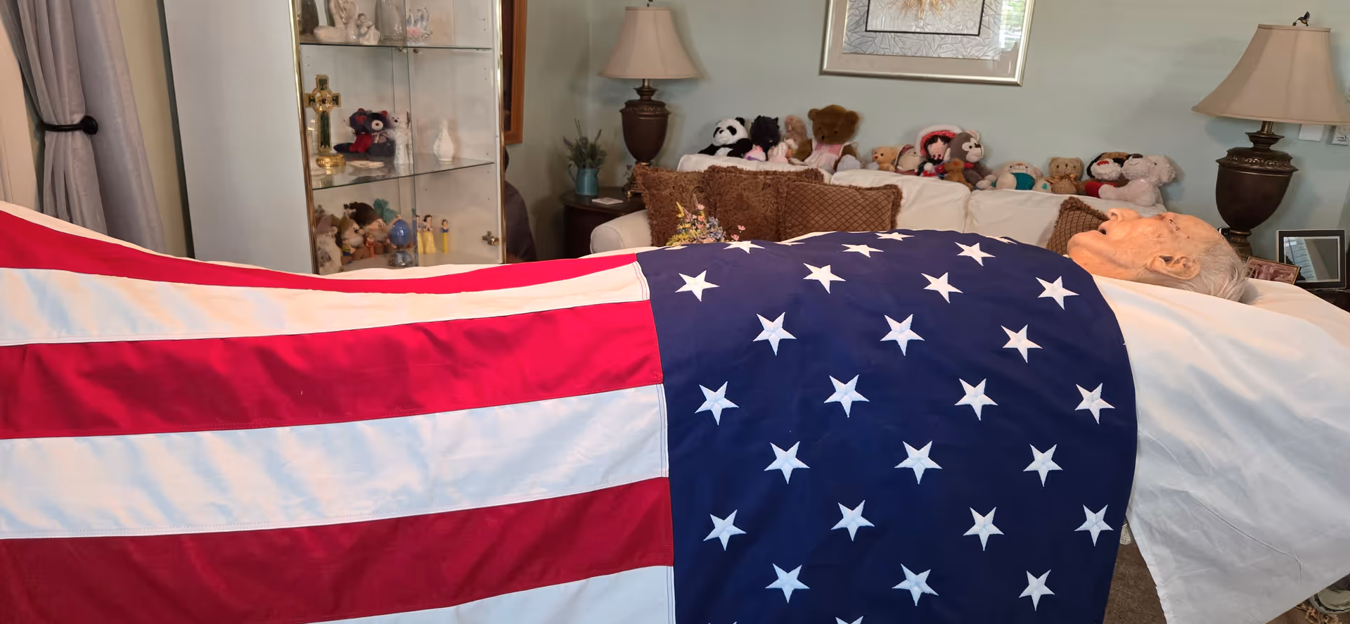 Elderly person lies in a bed draped with an American flag in a furnished residential room.