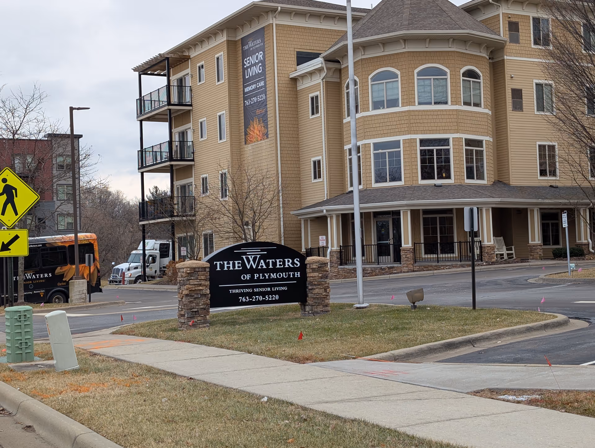 Front exterior of The Waters of Plymouth senior living building showing the entrance, landscaped sign, and driveway.