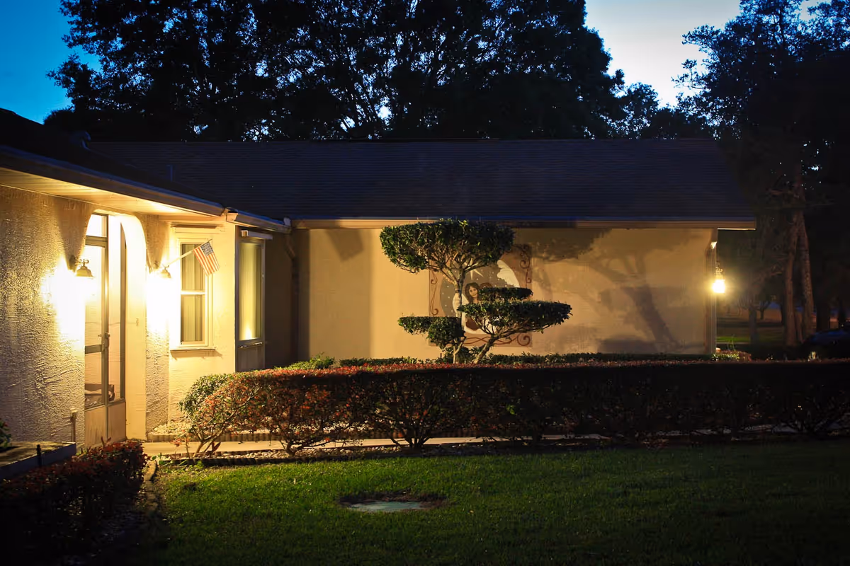 Exterior view of a single-story building at dusk with outdoor lights illuminating the entrance and a small American flag mounted near the door. There are neatly trimmed bushes and a uniquely shaped tree in front of the building, with a grassy lawn in the foreground and tall trees in the background.