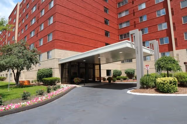 Red brick senior living building with a glass entrance canopy and landscaped driveway.