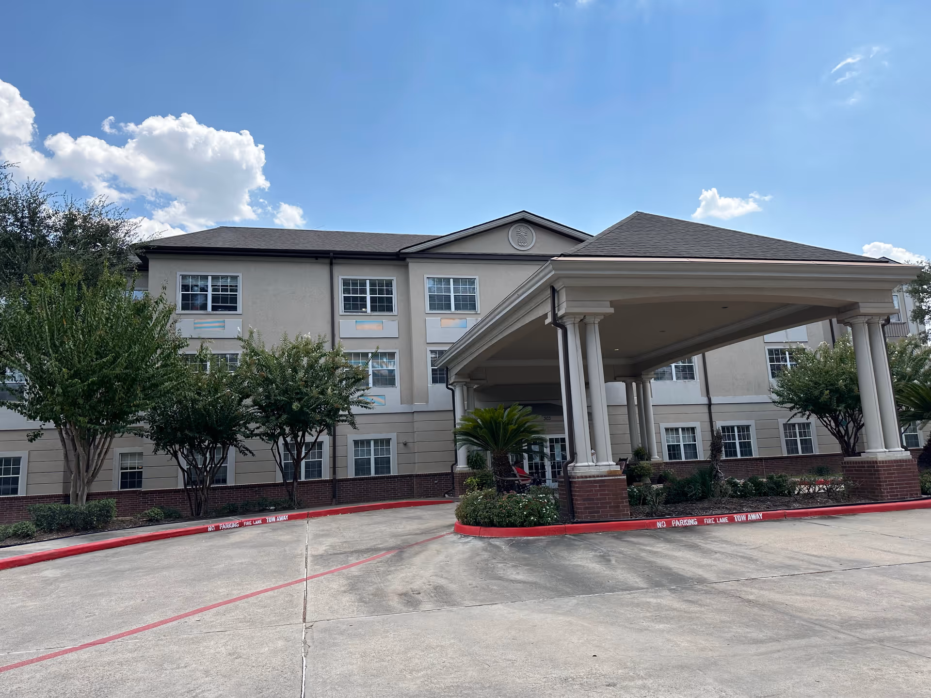 Three-story beige senior living building with a covered entrance and landscaped front, blue sky above.