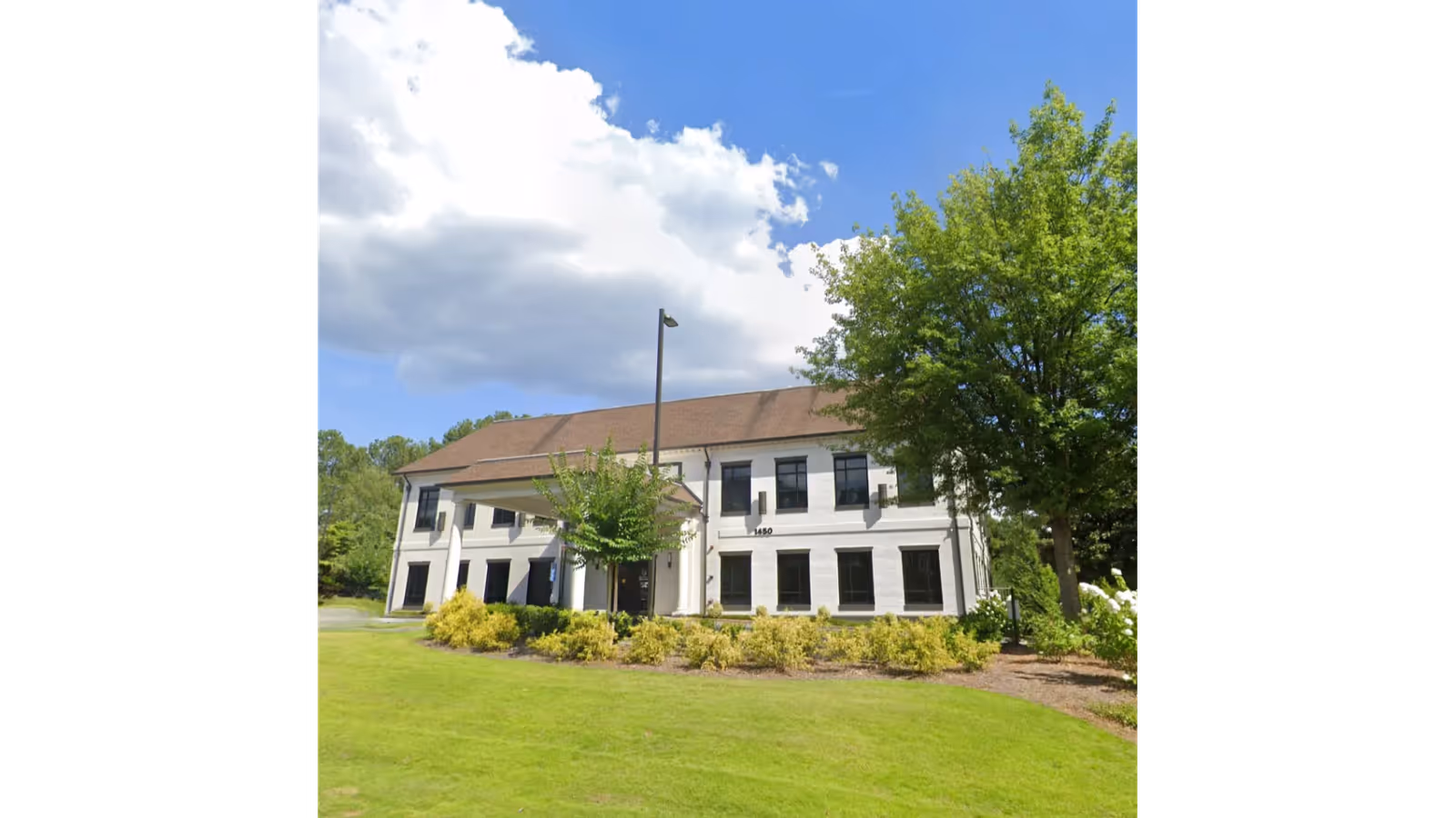 White two-story building with black-trim windows, surrounded by trees and manicured shrubbery.