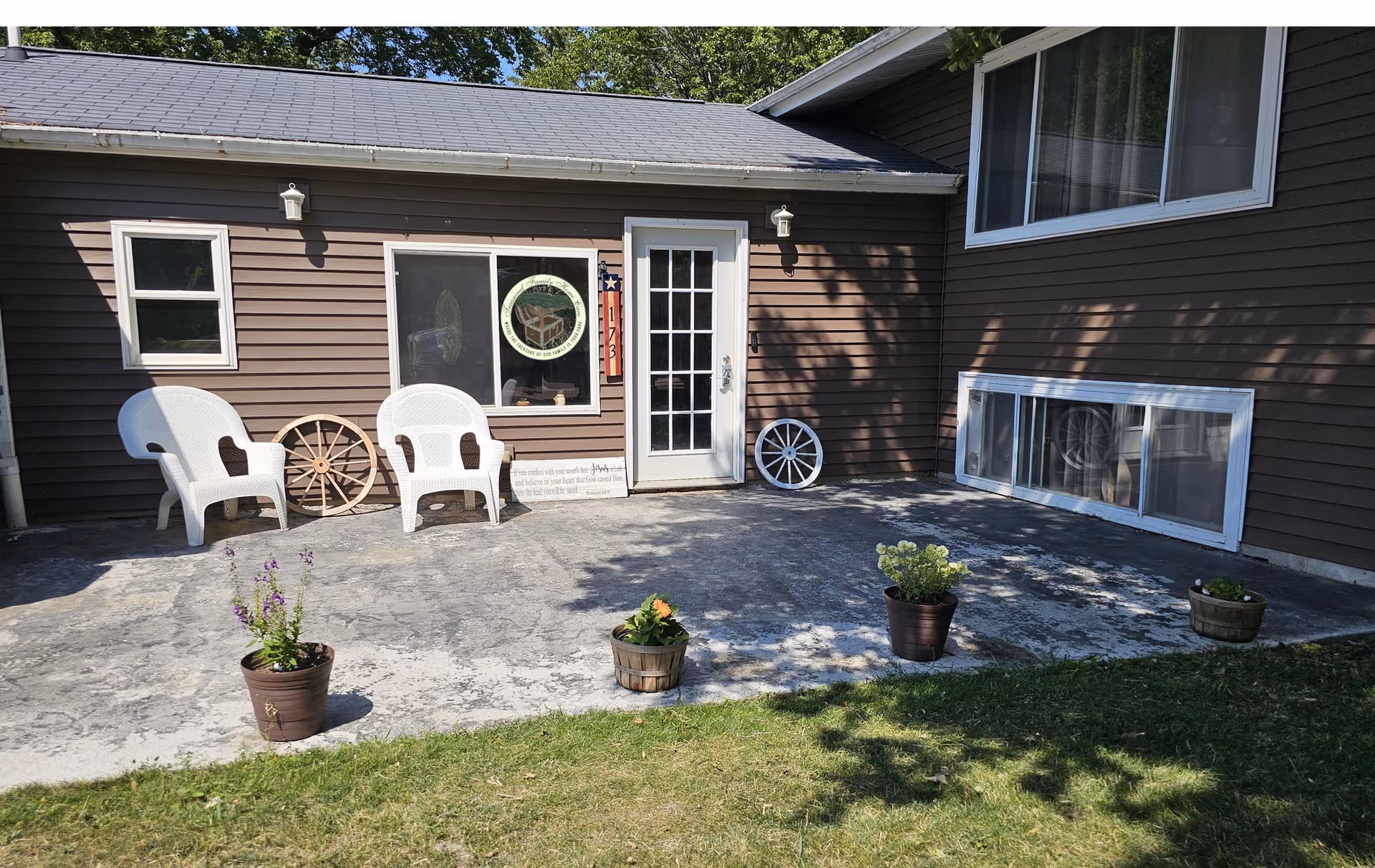 Brown single-story building exterior with a concrete patio, white chairs, and potted plants.