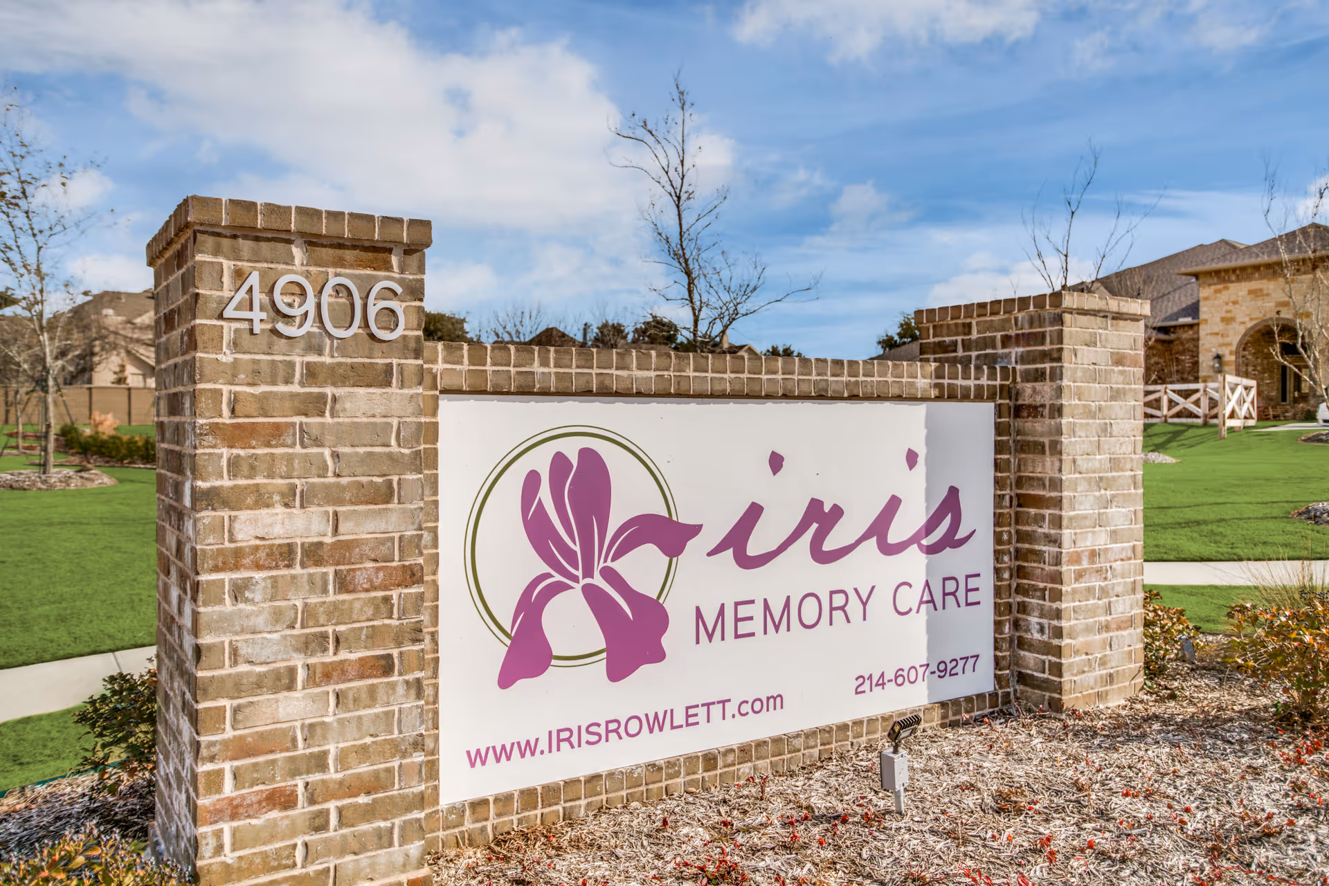 Brick entrance sign shows Iris Memory Care with a purple iris logo beside a grassy entrance.