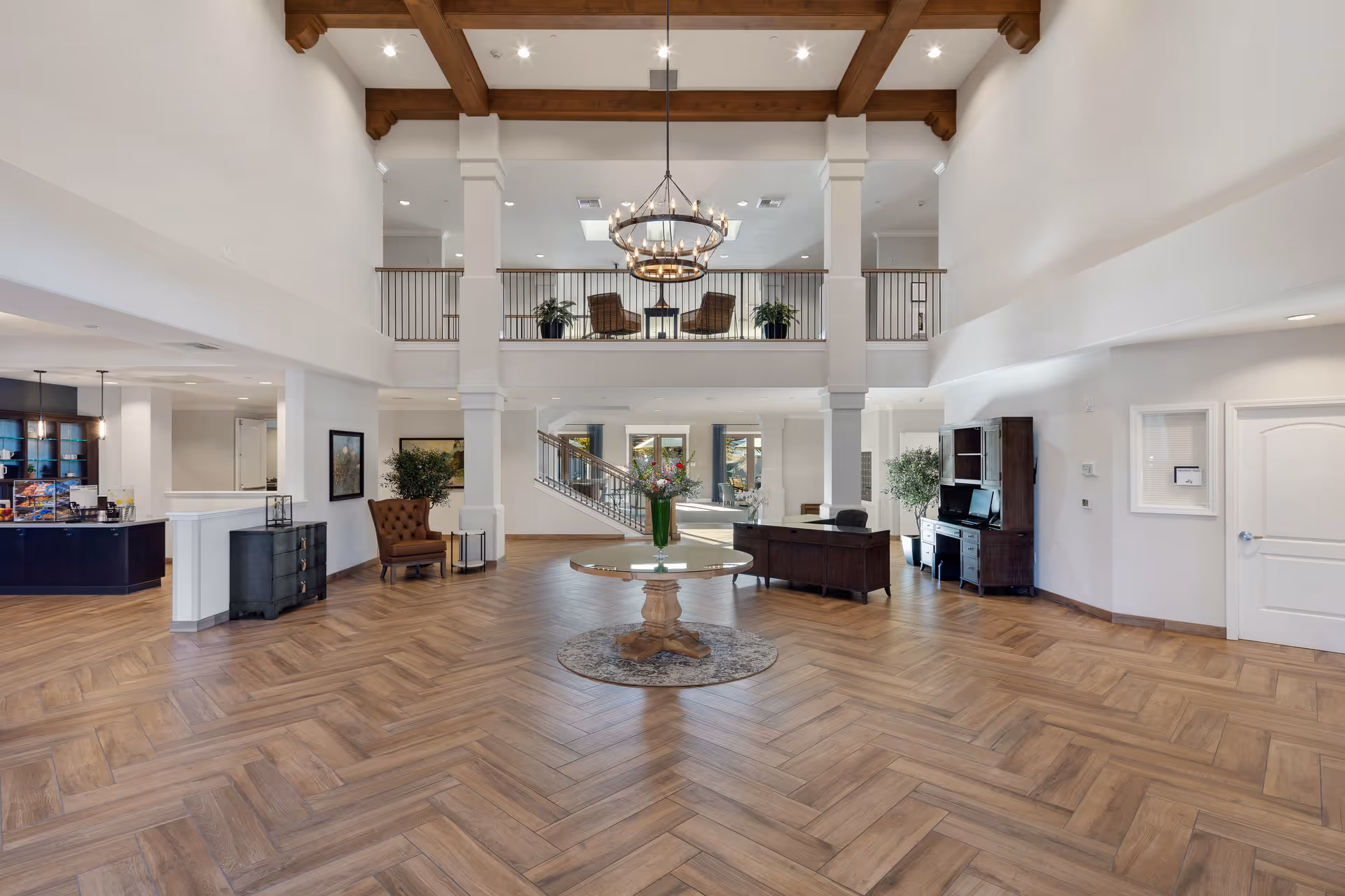 Spacious and elegant senior living facility lobby with high ceilings featuring wooden beams, a large circular chandelier, and a central round table with a flower vase. The room has wood-patterned flooring, a reception desk, comfortable seating areas, and a staircase leading to an upper balcony with additional seating and plants.
