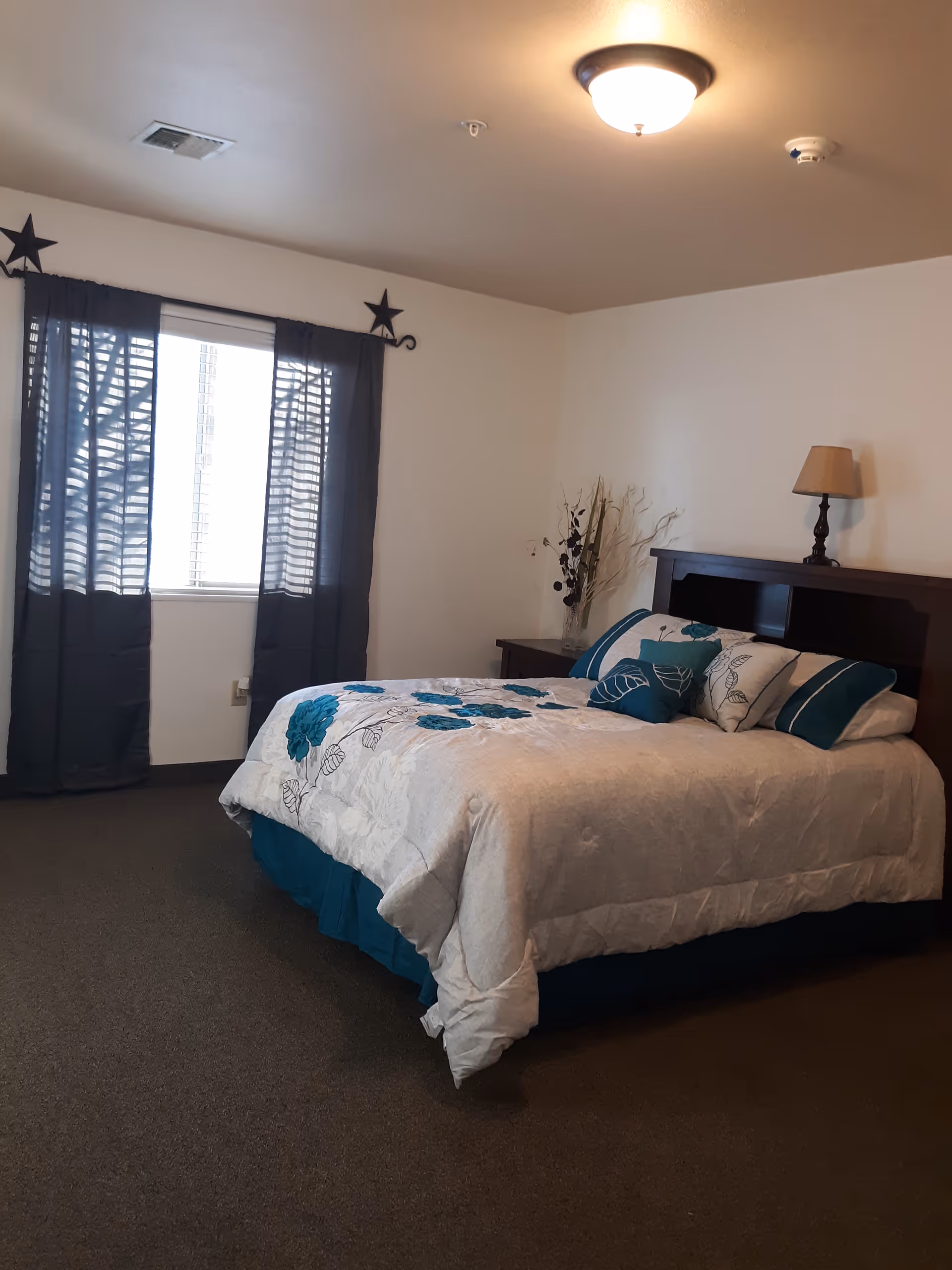 Neatly made bedroom with a large bed, nightstand, lamp, and dark curtains.